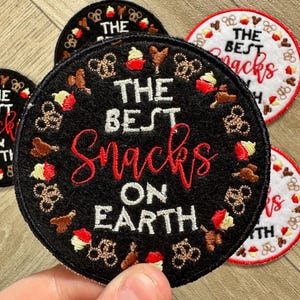 May include: Black embroidered patch with white text that reads "The Best Snacks On Earth". The patch is surrounded by a circle of embroidered food icons, including pretzels, Mickey Mouse ears, churros, and ice cream.