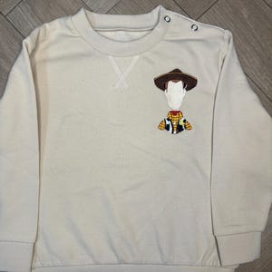 以下が含まれることがあります： トイストーリーのウッディが刺繍されたベージュのトレーナー。トレーナーは丸首で長袖です。