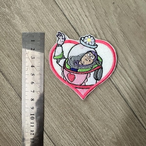 以下が含まれることがあります： ピンクのハートの中に漫画の宇宙飛行士が描かれた刺繍パッチ。宇宙飛行士は宇宙服と花の帽子をかぶっています。パッチの幅は約5cm、高さは約6.3cmです。