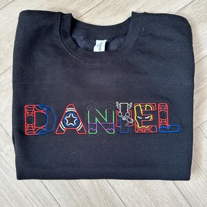 Puede incluir: Sudadera negra de cuello redondo con el nombre "DANIEL" bordado en letras coloridas con tem&aacute;tica de superh&eacute;roes. Las letras presentan dise&ntilde;os como una telara&ntilde;a, una estrella y rayos. La sudadera est&aacute; hecha de un material suave.