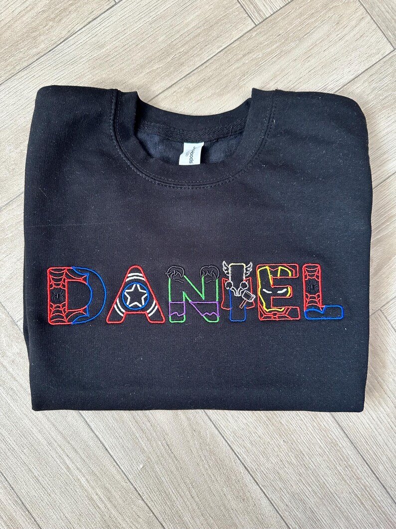 Puede incluir: Sudadera negra de cuello redondo con el nombre "DANIEL" bordado en letras coloridas con tem&aacute;tica de superh&eacute;roes. Las letras presentan dise&ntilde;os como una telara&ntilde;a, una estrella y rayos. La sudadera est&aacute; hecha de un material suave.