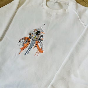 Puede incluir: Sudadera blanca con una imagen bordada de un droide de Star Wars. El droide es blanco, naranja y gris con un ojo negro.
