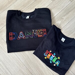 Puede incluir: Dos sudaderas negras. Una presenta el nombre "DANIEL" bordado en letras coloridas con tem&aacute;tica de superh&eacute;roes. La otra tiene un dise&ntilde;o de conejitos superh&eacute;roes coloridos. Ambas est&aacute;n planas sobre una superficie de madera.