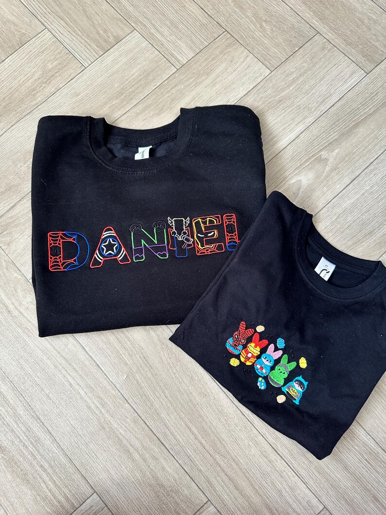 Puede incluir: Dos sudaderas negras. Una presenta el nombre "DANIEL" bordado en letras coloridas con tem&aacute;tica de superh&eacute;roes. La otra tiene un dise&ntilde;o de conejitos superh&eacute;roes coloridos. Ambas est&aacute;n planas sobre una superficie de madera.