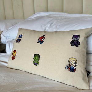 Peut inclure: Un oreiller rectangulaire blanc cassé brodé de personnages de dessins animés colorés. Les personnages incluent Captain America, Spider-Man, Black Panther, Iron Man, Hulk et Thor. L'oreiller est posé sur un lit blanc.