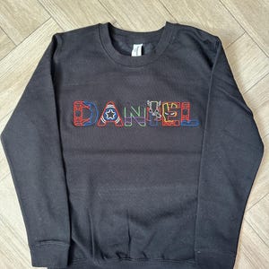 Puede incluir: Sudadera negra de cuello redondo con el nombre "DANIEL" bordado en letras coloridas con tem&aacute;tica de superh&eacute;roes. Las letras presentan dise&ntilde;os como una telara&ntilde;a, una estrella y rayos. La sudadera tiene mangas largas y un dobladillo acanalado.