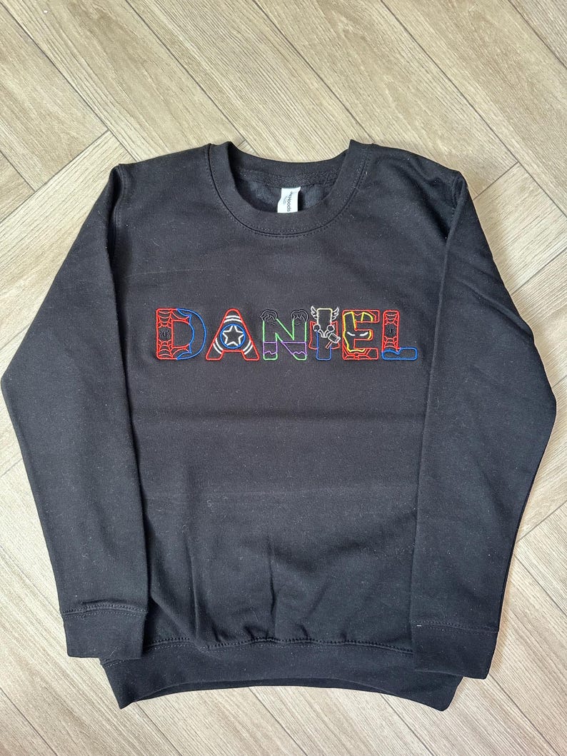 Puede incluir: Sudadera negra de cuello redondo con el nombre "DANIEL" bordado en letras coloridas con tem&aacute;tica de superh&eacute;roes. Las letras presentan dise&ntilde;os como una telara&ntilde;a, una estrella y rayos. La sudadera tiene mangas largas y un dobladillo acanalado.