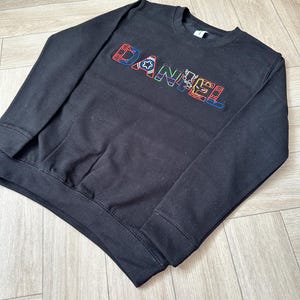 Puede incluir: Sudadera negra de cuello redondo con el nombre "DANIEL" bordado en letras coloridas. Las letras est&aacute;n delineadas en varios colores, incluyendo rojo, azul, verde y amarillo. La sudadera tiene dobladillo y pu&ntilde;os acanalados.