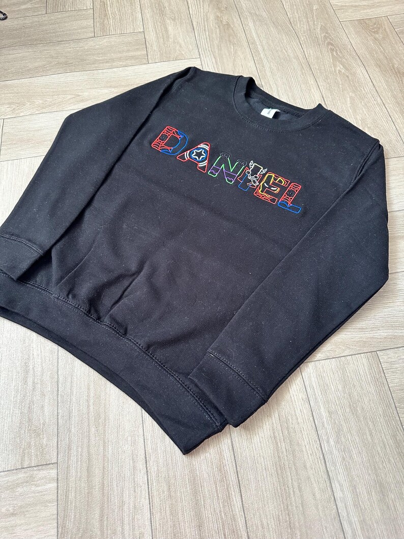 Puede incluir: Sudadera negra de cuello redondo con el nombre "DANIEL" bordado en letras coloridas. Las letras est&aacute;n delineadas en varios colores, incluyendo rojo, azul, verde y amarillo. La sudadera tiene dobladillo y pu&ntilde;os acanalados.