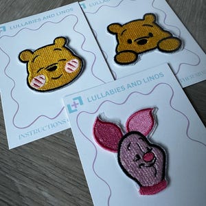 Peut inclure: Trois écussons brodés représentant des personnages de dessins animés. Deux présentent un ours jaune avec des contours noirs, et le troisième un cochon rose avec de grandes oreilles. Chaque écusson est sur une carte blanche avec le texte "Lullabies and Linos".
