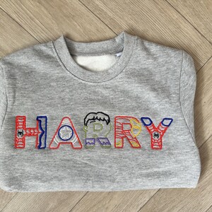 Puede incluir: Una sudadera gris con el nombre "HARRY" bordado en letras coloridas con tem&aacute;tica de superh&eacute;roes. Las letras est&aacute;n delineadas en rojo, azul y verde, con detalles de personajes.