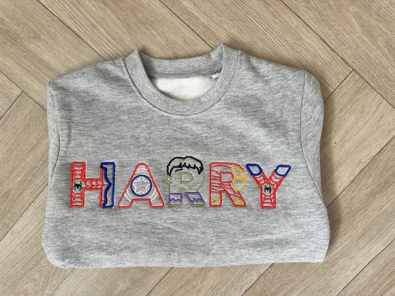 Puede incluir: Una sudadera gris con el nombre "HARRY" bordado en letras coloridas con tem&aacute;tica de superh&eacute;roes. Las letras est&aacute;n delineadas en rojo, azul y verde, con detalles de personajes.