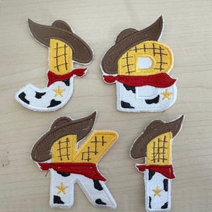 Könnte beinhalten: Vier gestickte Aufnäher in Form der Buchstaben J, B, K und I. Jeder Buchstabe hat einen Cowboyhut, ein rotes Halstuch und ein Kuhmuster. Die Buchstaben sind gelb mit braunen Umrissen und einem goldenen Stern.