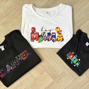 Puede incluir: Tres camisetas con dise&ntilde;os bordados. Una camiseta blanca con las palabras "boy mama" y personajes de superh&eacute;roes. Una camiseta negra con el nombre "DANIEL" en una fuente de superh&eacute;roe. Otra camiseta negra con un grupo de personajes de superh&eacute;roes.