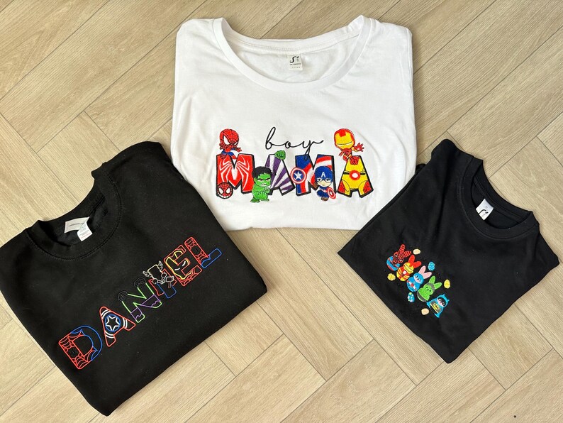 Puede incluir: Tres camisetas con dise&ntilde;os bordados. Una camiseta blanca con las palabras "boy mama" y personajes de superh&eacute;roes. Una camiseta negra con el nombre "DANIEL" en una fuente de superh&eacute;roe. Otra camiseta negra con un grupo de personajes de superh&eacute;roes.