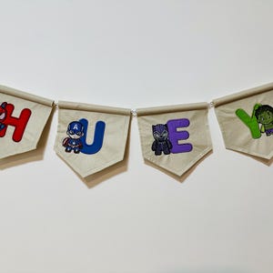 May include: A decorative banner spelling "HEY" with superhero characters. Each beige pennant displays a letter and a superhero: Spider-Man, Captain America, Black Panther, and the Hulk. The letters are in red, blue, and purple.