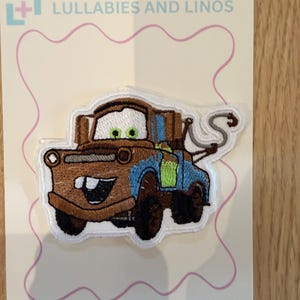 Op de afbeelding: Een geborduurde patch met een bruine en blauwe takelwagen-personage met groene ogen en een witte sleephaak. De patch is bevestigd op een witte displaykaart met de tekst LULLABIES AND LINOS bovenaan en INSTRUCTIONS ON OTHER SIDE onderaan.
