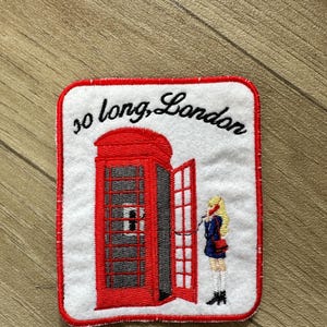 May include: Embroidered patch featuring a red London telephone booth with an open door and a person on the phone. The patch has the words "so long, London" in cursive script.