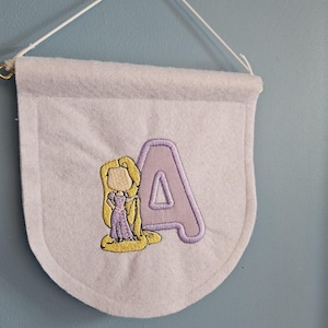 Puede incluir: Un banderín de fieltro morado claro con un anillo dorado y una cuerda blanca para colgar. El banderín presenta una letra "A" bordada y una imagen de dibujos animados de una princesa con un vestido morado y pelo largo rubio.