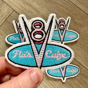 May include: Five embroidered iron-on patches with a blue and white oval design. The oval has the text "Fla's Cafe" in pink lettering. The centre of the patch features a pink and white stylised "V8" logo.