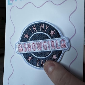 May include: Embroidered patch with the text "IN MY SHOWGIRL ERA" in pink and white on a navy blue background. The patch is circular with pink star accents and a light blue border. The patch is on a white card with the text "Lullabies and Linos".