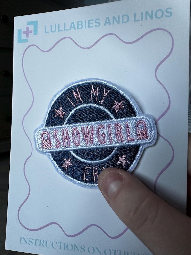 Puede incluir: Parche bordado con el texto "IN MY SHOWGIRL ERA" en rosa y blanco sobre fondo azul marino. El parche es circular con detalles de estrellas rosas y un borde azul claro. El parche est&aacute; sobre una tarjeta blanca con el texto "Lullabies and Linos".