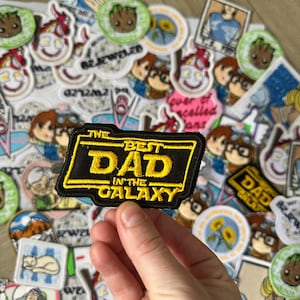 Peut inclure: Un écusson noir et jaune avec le texte "THE BEST DAD IN THE GALAXY" est tenu dans une main. L'écusson est entouré de divers écussons brodés représentant des personnages de dessins animés et d'autres motifs.
