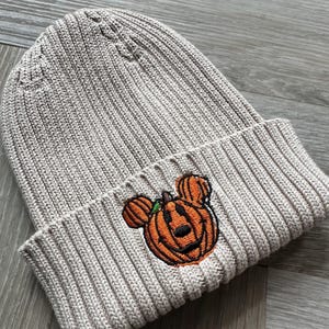 Op de afbeelding: Een beige geribbelde gebreide beanie met een geborduurd pompoenontwerp met Mickey Mouse oren. De pompoen is oranje met een zwart gezicht en groene stengel. Een herfstaccessoire.