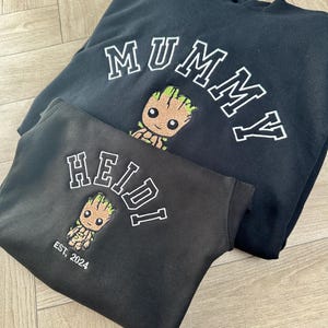 May include: Two black hooded sweatshirts with embroidered designs. The larger sweatshirt says "MUMMY" in white letters with a green and brown cartoon character. The smaller sweatshirt says "HEIDI" in white letters with a green and brown cartoon character and "EST. 2024" in white letters.