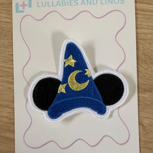 May include: An embroidered patch featuring a blue wizard hat with gold stars and a crescent moon, flanked by two black circular ears. The patch is mounted on a white display card with the text LULLABIES AND LINOS and INSTRUCTIONS ON OTHER SIDE.
