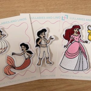 Puede incluir: Una colección de parches bordados con personajes animados que incluyen una sirena, una figura con traje blanco y una princesa con vestido rosa, presentados en tarjetas de soporte blancas etiquetadas Lullabies and Linos con instrucciones.