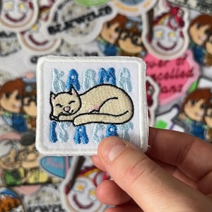 Könnte beinhalten: Ein weißes quadratisches Patch mit dem Text "Karma is a Cat" in blauen Buchstaben. Das Patch zeigt eine beige Katze, die sich in Schlafposition zusammenrollt. Das Patch ist von anderen bunten Patches umgeben.