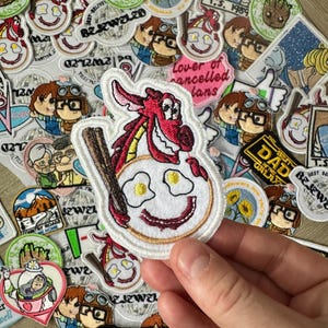May include: A selection of embroidered patches with diverse designs. One patch depicts a red dragon with a baseball bat over a smiling face with fried eggs. Other patches feature cartoon characters, text, and floral motifs.