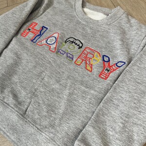 Puede incluir: Una sudadera gris con el nombre "HARRY" bordado en letras coloridas. Cada letra est&aacute; dise&ntilde;ada con un tema de superh&eacute;roe, con detalles en rojo, azul, amarillo y verde. La sudadera tiene cuello redondo y pu&ntilde;os acanalados.
