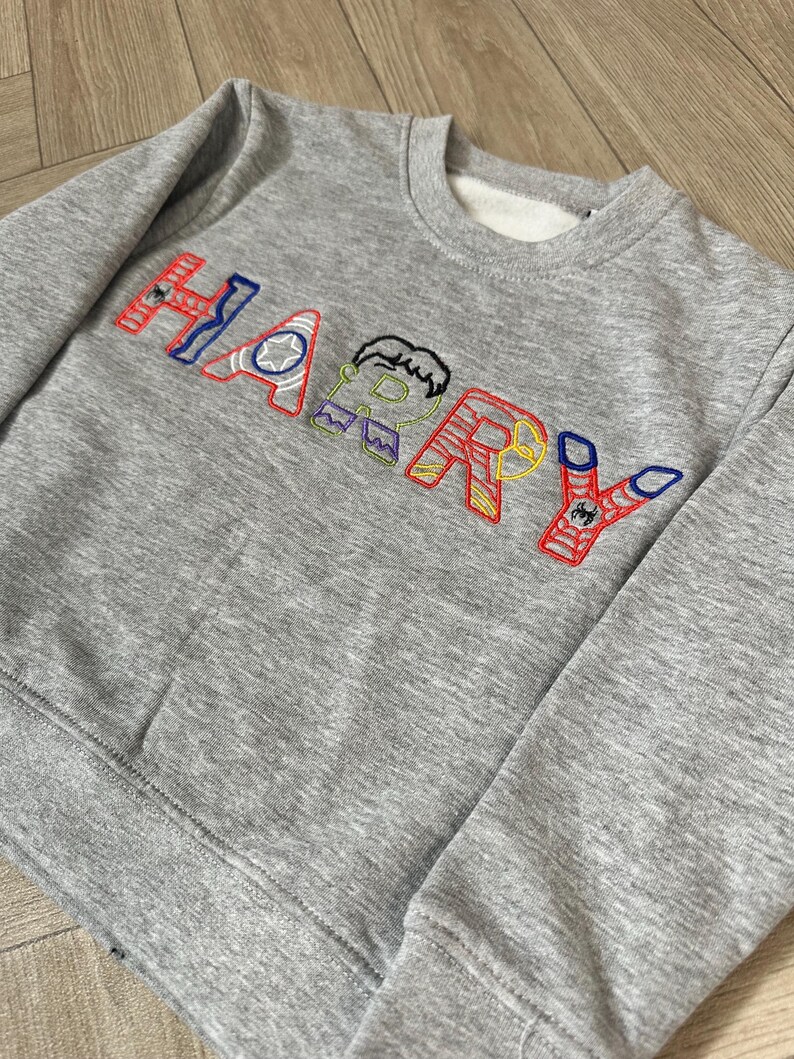 Puede incluir: Una sudadera gris con el nombre "HARRY" bordado en letras coloridas. Cada letra est&aacute; dise&ntilde;ada con un tema de superh&eacute;roe, con detalles en rojo, azul, amarillo y verde. La sudadera tiene cuello redondo y pu&ntilde;os acanalados.
