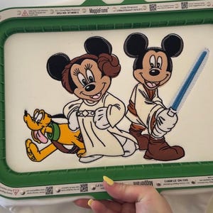 May include: An embroidery hoop featuring Mickey Mouse and Minnie Mouse dressed as Jedi characters, with Pluto. The design is set against a white background, framed by a green border. The hoop measures approximately 7.7 x 12.5 inches.