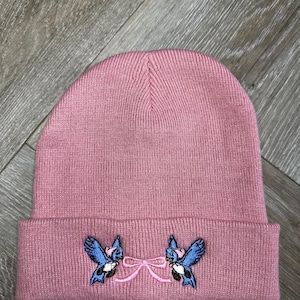 May include: A dusky pink beanie hat with a turned-up brim. It has an embroidered design of two blue birds facing each other, linked by a pink bow. The hat is made of a ribbed knit fabric.