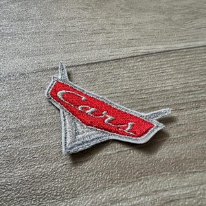 May include: Embroidered patch with the word "Cars" in white script on a red rectangular background. The patch has a silver border and a stylized design. Suitable for clothing or accessories.