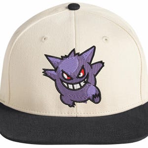 Könnte beinhalten: Eine cremefarbene Baseballkappe mit schwarzem Schirm und einem kleinen schwarzen Knopf an der Oberseite. Auf der Vorderseite befindet sich eine Stickerei des violetten Gengar-Charakters mit roten Augen und einem breiten, gezahnten Grinsen.