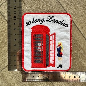 May include: Embroidered patch featuring a red London telephone booth with an open door and a person standing nearby. The text "so long, London" is written above the booth in black script. The patch has a white background and a red border.