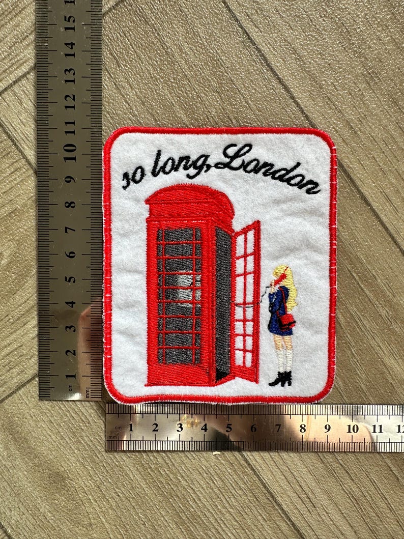 May include: Embroidered patch featuring a red London telephone booth with an open door and a person standing nearby. The text "so long, London" is written above the booth in black script. The patch has a white background and a red border.