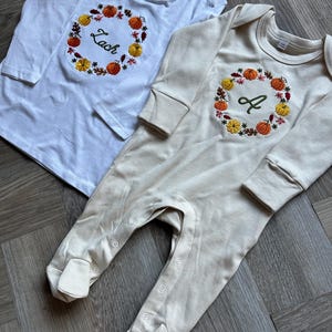 May include: Two baby outfits: a white long-sleeve shirt and a beige footed onesie. Both feature a fall-themed embroidered wreath with pumpkins and leaves. The shirt has the name "Zach", and the onesie has the letter "A".