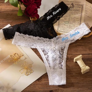 May include: Black and white lace thong underwear. The black thong has the name "Mrs. Rivera" embroidered on the waistband. The white thong has the name "Mrs. Smith" embroidered on the waistband. Both thongs have a delicate lace design.