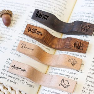 May include: Wooden book page holders with engraved names and symbols. The holders are in various wood tones, including dark brown, medium brown, and light wood. Each holder has a unique design, such as a butterfly, book, brain, and moon with stars.