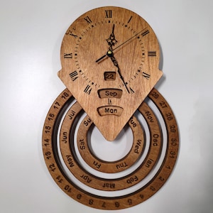 Peut inclure: Horloge murale en bois au design unique. L'horloge affiche des chiffres romains, une date et des anneaux rotatifs pour le mois et le jour. Fabriquée en bois brun clair, elle présente une esthétique moderne. Dimensions en centimètres.