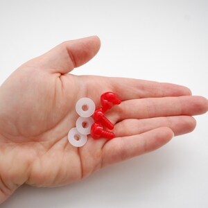 May include: A hand holding a collection of small craft supplies. The items include three white, circular washers and several red, plastic, screw-shaped components. The objects are set against a plain white background.