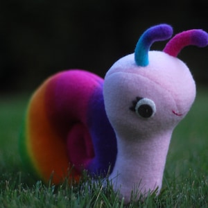 Op de afbeelding: Een pluche slak speelgoed met een roze lichaam en een regenboogkleurige schelp. De slak heeft twee gebogen antennes met paarse, blauwe en roze uiteinden. Het heeft een knoopoog en een gestikte glimlach, tegen een achtergrond van groen gras.