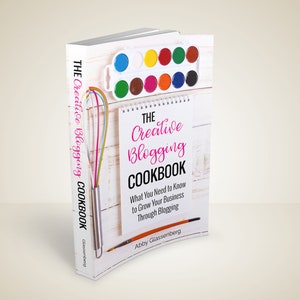May include: A book titled "The Creative Blogging Cookbook" with a watercolor paint set and a whisk on top. The book's cover features the title in pink script and the subtitle "What You Need to Know to Grow Your Business Through Blogging."