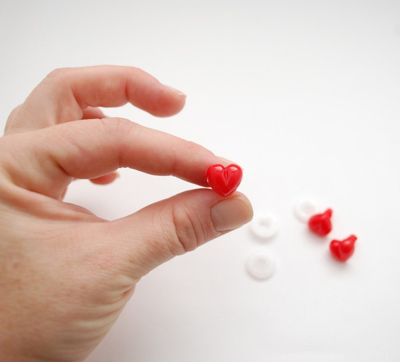 May include: A small, glossy red heart-shaped bead held between fingers. Several other red heart beads and white circular beads are scattered on a white surface. The heart bead is the focal point.