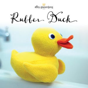 May include: A plush yellow rubber duck with an orange beak and blue eyes. The duck is sitting on a white surface. The text "Rubber Duck" is written in cursive above the duck.